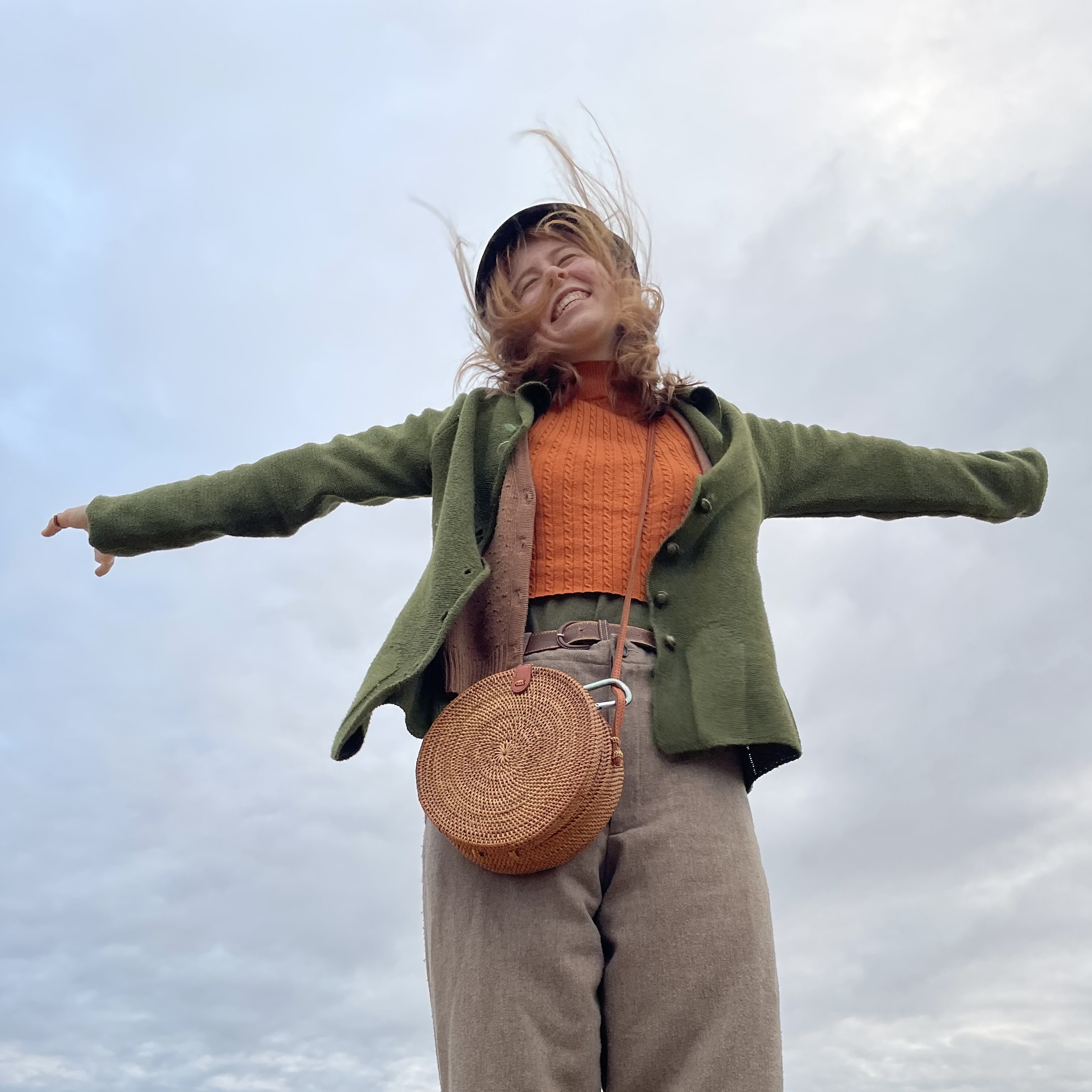Woman with outstretched arms, smiling, wearing a green jacket and orange top against a cloudy sky.